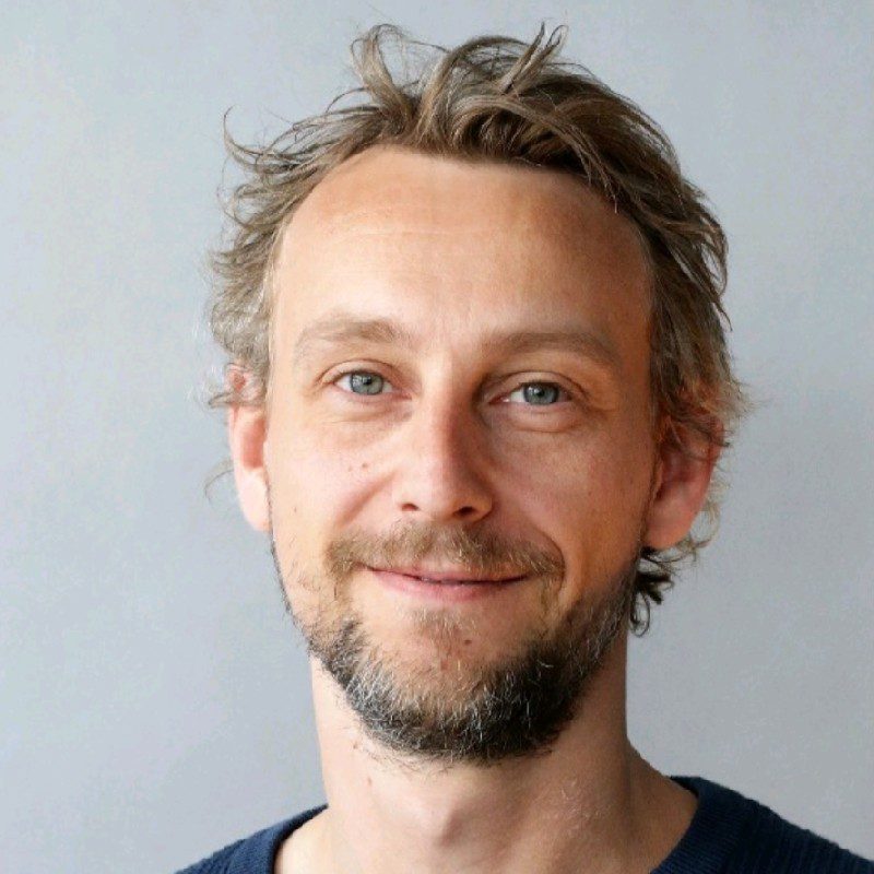 a man with tousled hair and a beard smiling softly at the camera wearing a dark sweater against a light gray background digital production A man with tousled hair and a beard smiling softly at the camera, wearing a dark sweater against a light gray background.
