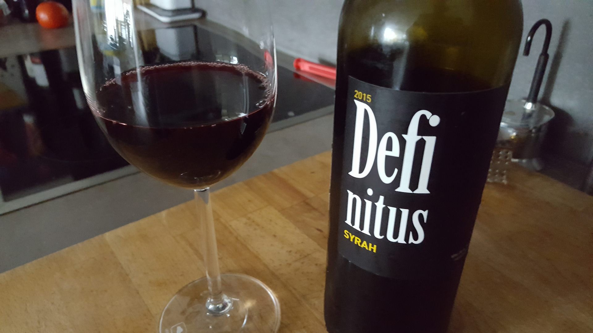 a glass of red wine filled to the rim next to a bottle of defi nitus syrah from 2015 placed on a wooden surface the glass reflects light showing rich deep hues of red while the bottle has a sleek black label with white lettering digital production A glass of red wine filled to the rim next to a bottle of 'Defi nitus' Syrah from 2015, placed on a wooden surface. The glass reflects light, showing rich, deep hues of red, while the bottle has a sleek black label with white lettering.