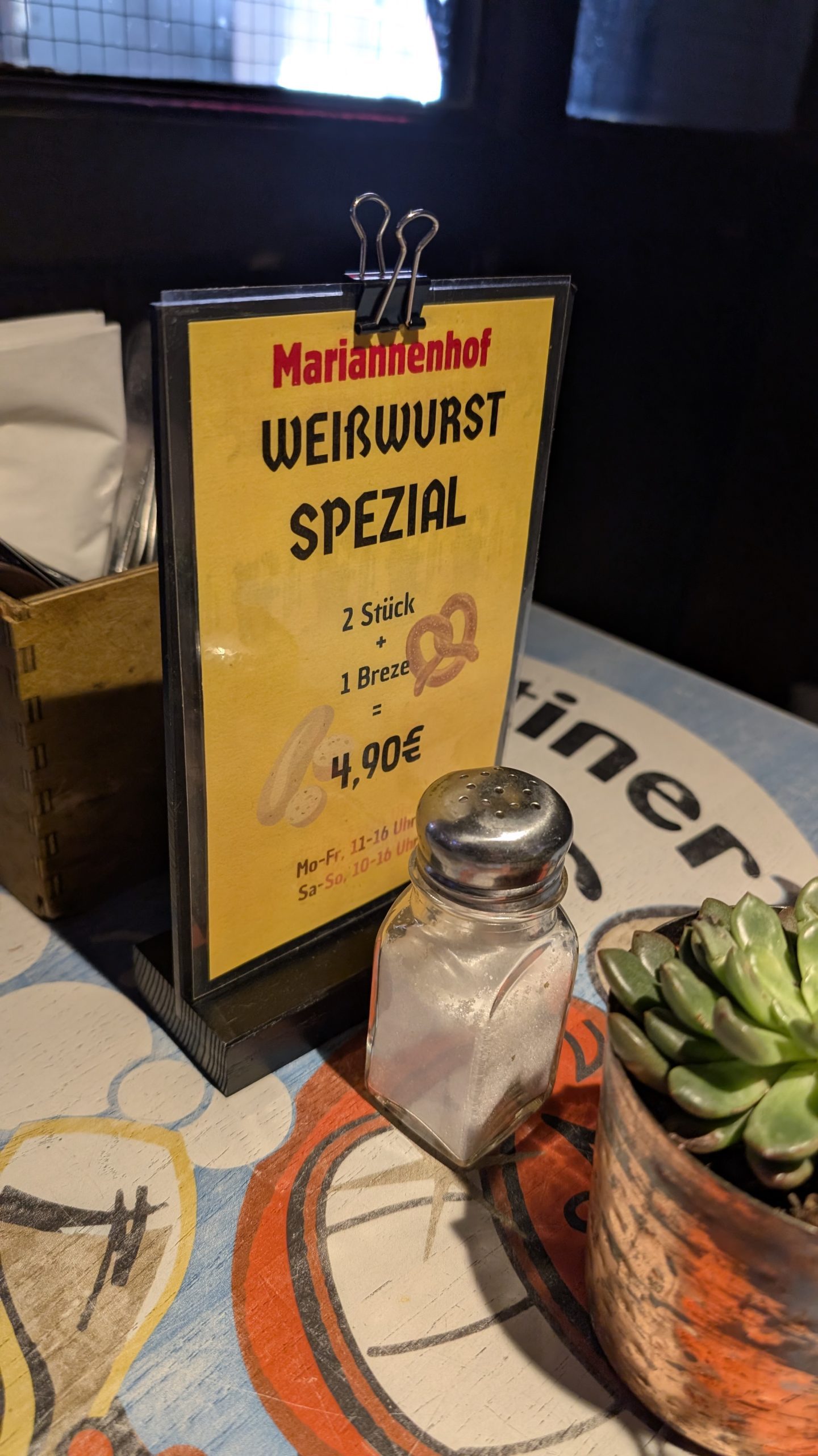 a menu card on a table featuring a special dish titled weißwurst spezial from marienhof the menu lists two sausages and a pretzel for €490 a salt shaker and a potted succulent are placed nearby digital production A menu card on a table featuring a special dish titled 'Weißwurst Spezial' from Marienhof. The menu lists two sausages and a pretzel for €4.90. A salt shaker and a potted succulent are placed nearby.