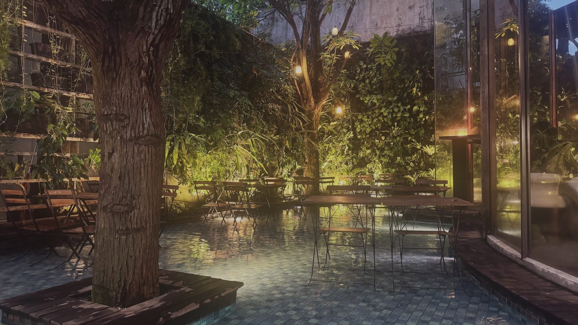 an indoor garden with a large tree illuminated by soft lights surrounding it are several tables and chairs placed on a tiled floor with greenery on the walls creating a tranquil atmosphere digital production An indoor garden with a large tree, illuminated by soft lights. Surrounding it are several tables and chairs placed on a tiled floor, with greenery on the walls, creating a tranquil atmosphere.