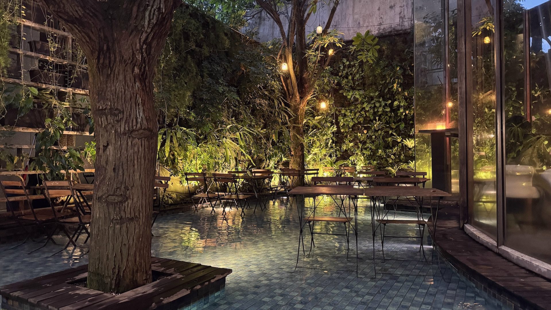 a tranquil outdoor seating area with wooden tables and chairs surrounded by greenery soft lighting illuminates the space reflecting off the water in the foreground digital production A tranquil outdoor seating area with wooden tables and chairs surrounded by greenery. Soft lighting illuminates the space, reflecting off the water in the foreground.