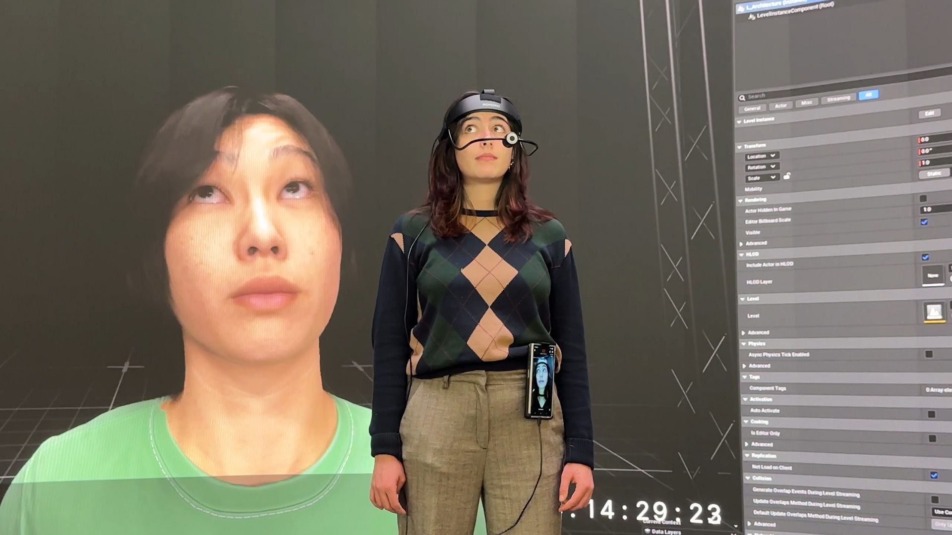 a person wearing a headset stands in front of a large digital screen displaying their own face in a 3d model the background features a grid and interface elements highlighting a tech setup digital production A person wearing a headset stands in front of a large digital screen displaying their own face in a 3D model. The background features a grid and interface elements, highlighting a tech setup.