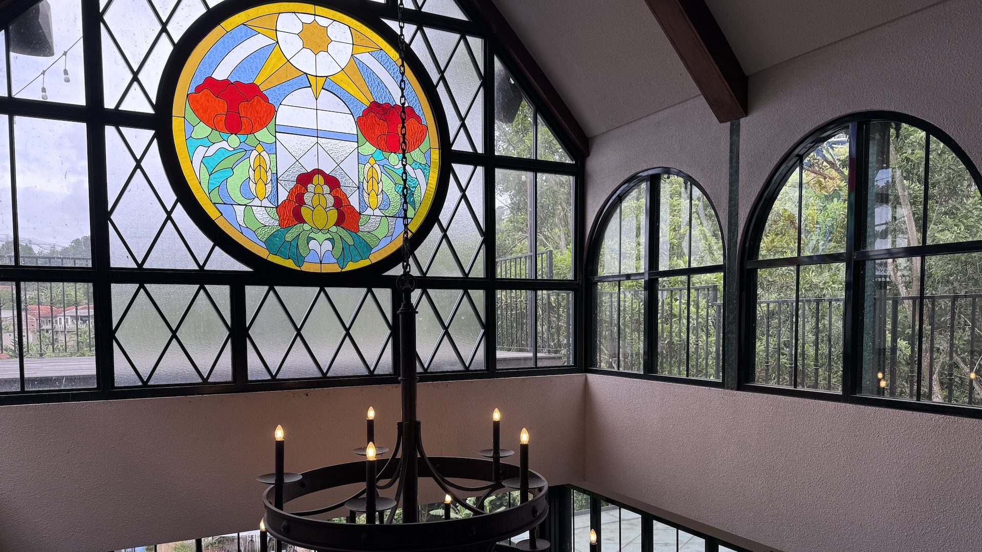 a large stained glass window featuring colorful flowers is displayed in a building with tall arched windows a chandelier with candles hangs below the window illuminating the interior space digital production A large stained glass window featuring colorful flowers is displayed in a building with tall, arched windows. A chandelier with candles hangs below the window, illuminating the interior space.